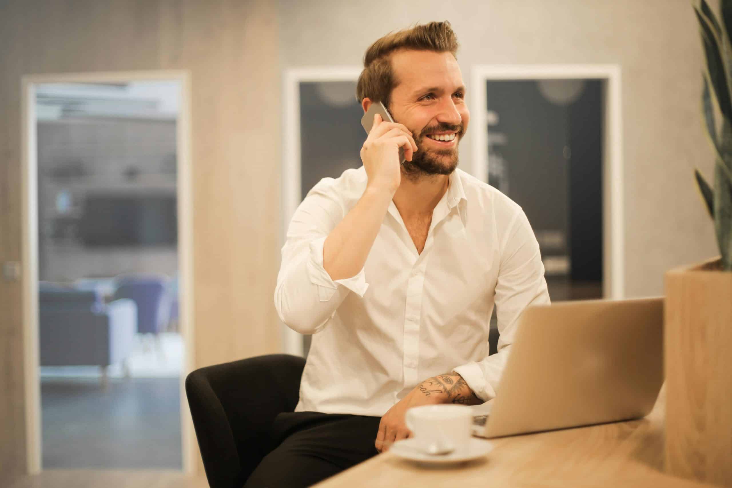 Homme souriant en chemise blanche parlant au téléphone, assis devant un ordinateur portable dans un bureau moderne.