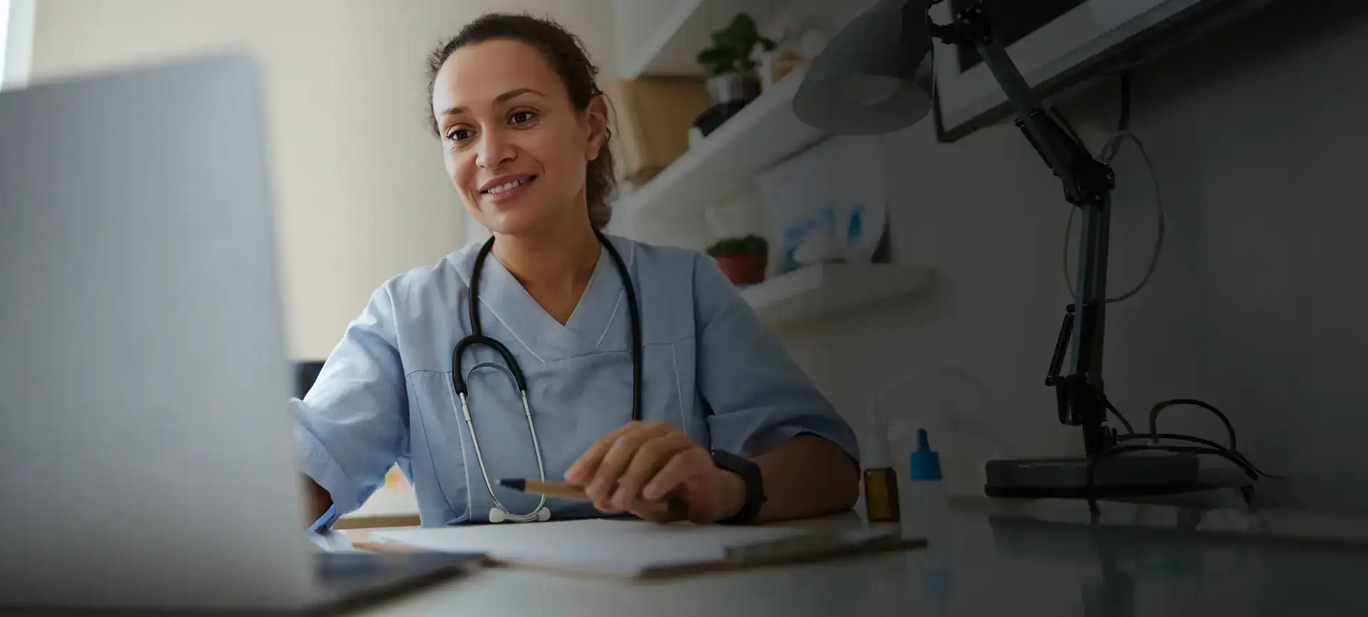 Female healthcare worker with stethoscope working on laptop in clinic.