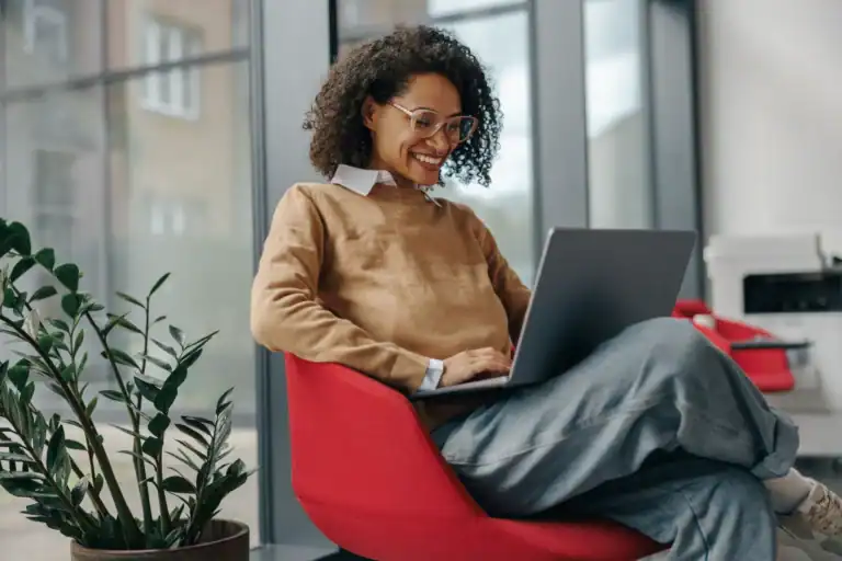 Happy woman using laptop in modern office with plants and large windows.