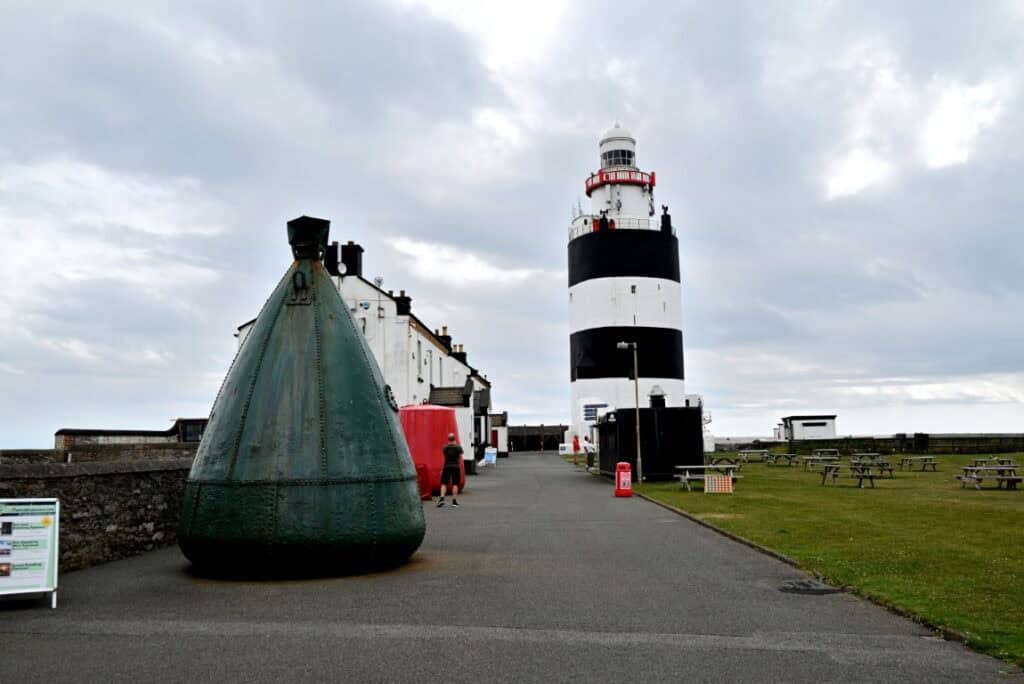 Lighthouse on the Irish Copper Coast with a large green buoy in the foreground and cloudy sky overhe.