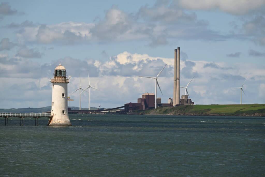 Lighthouse along the Irish coast with wind turbines and industrial buildings in the background.