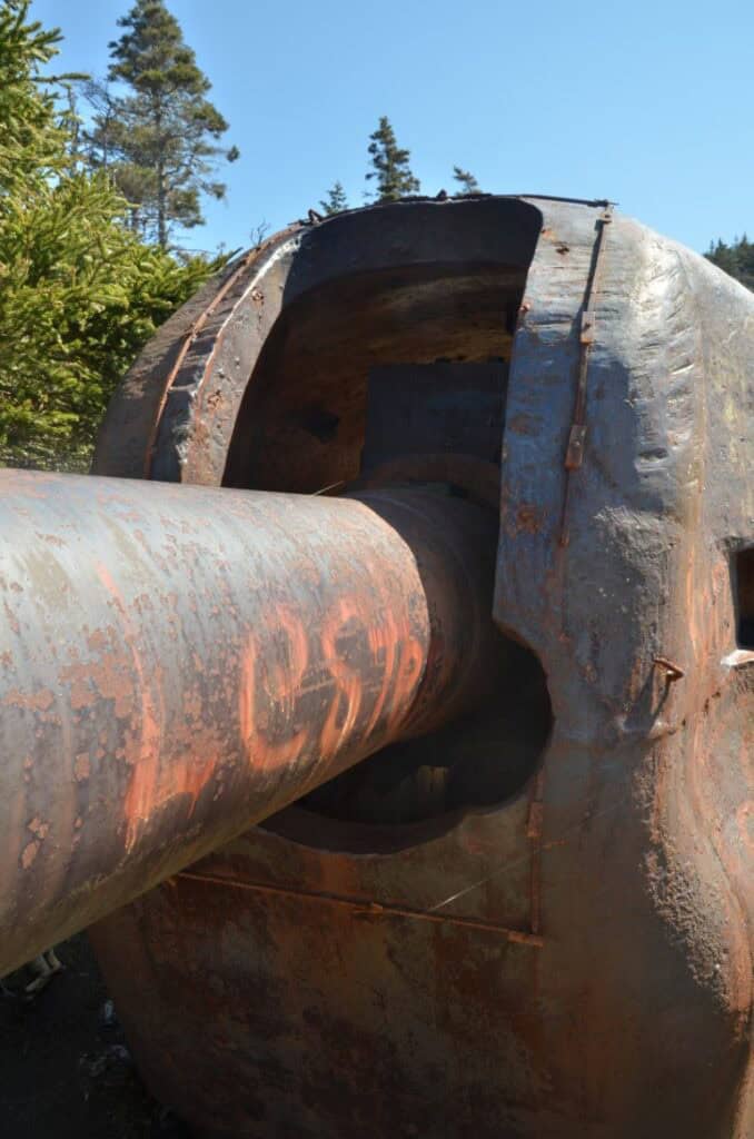 Rusty submarine hull with large periscope structure, surrounded by trees and clear blue sky.