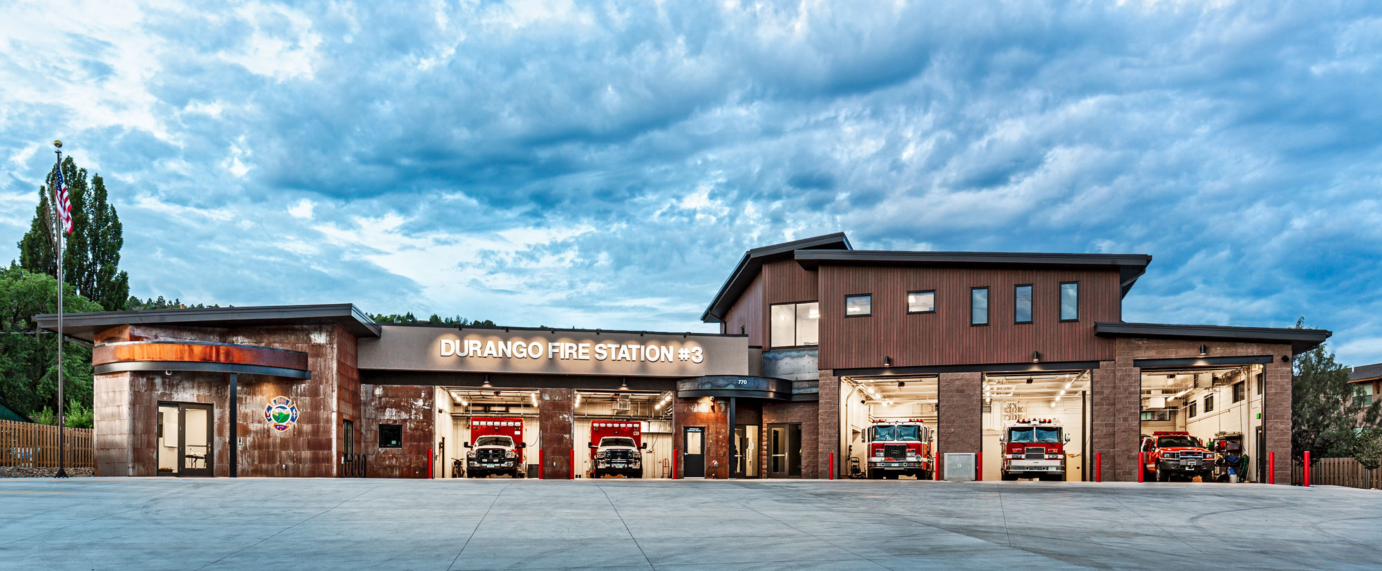 Modern fire station building with multiple garage bays and fire trucks under a cloudy sky.