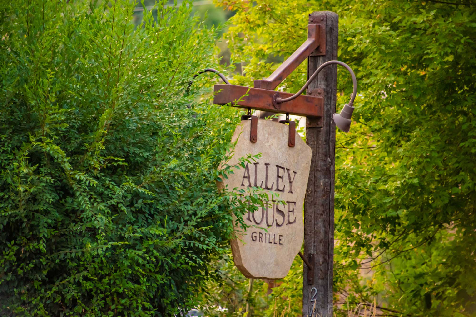 Alley House Grille outdoor sign on a rustic wooden post surrounded by greenery.