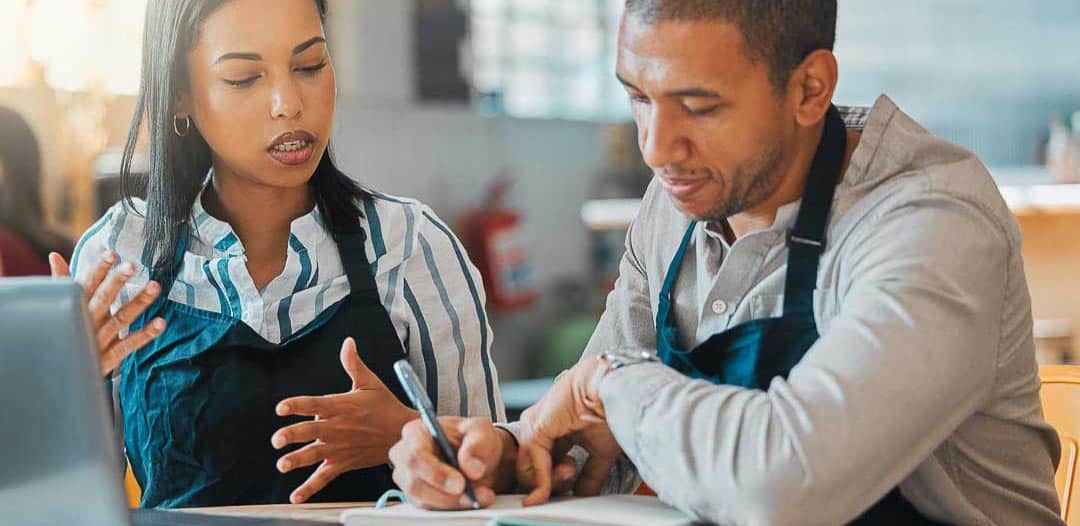 Restaurant owners in aprons reviewing notes and financial plans together with a laptop.