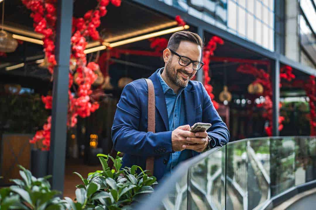 Male entrepreneur smiling while checking his phone outside a London venue decorated with red flowers.