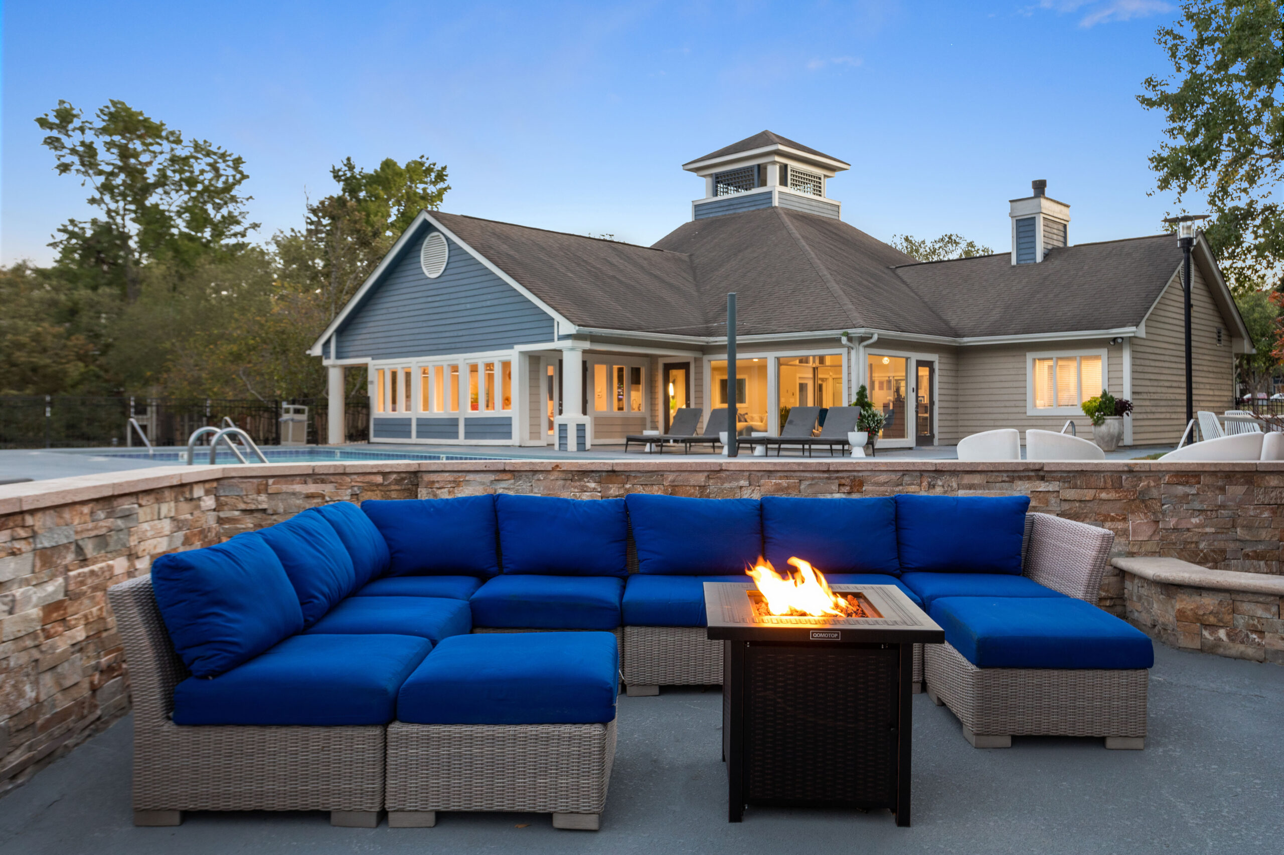 Outdoor sectional lounge sofa with blue cushions and firepit burning at dusk. The clubhouse is in the background.