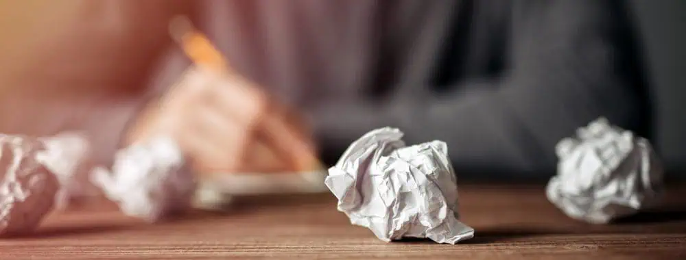 A man trying to write on a pad of papper, surrounded by balls of screwed up paper from previous failures