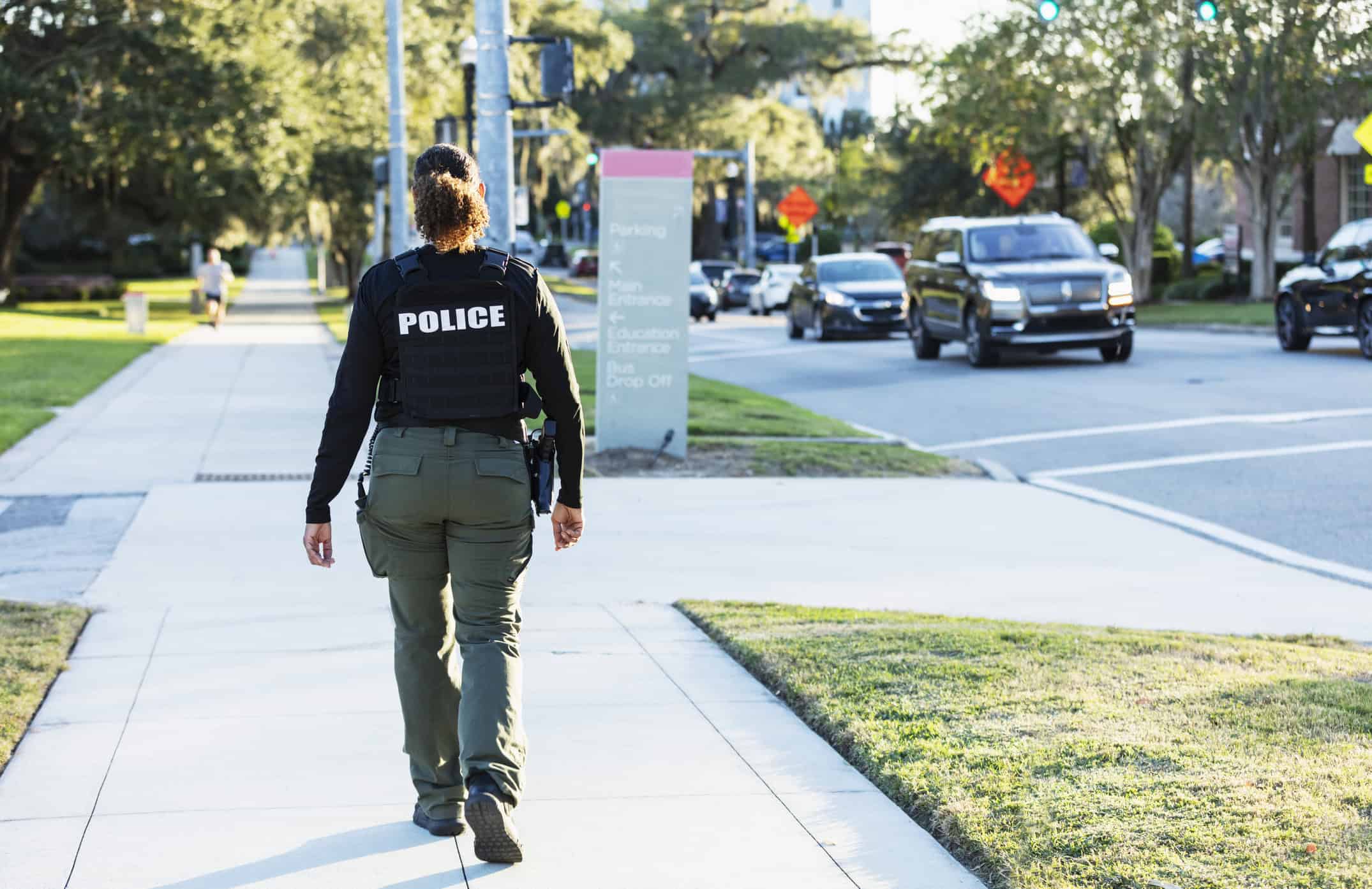 Rear view of a mature multiracial policewoman patrolling an urban area on foot. She is walking on a sidewalk away from the camera.