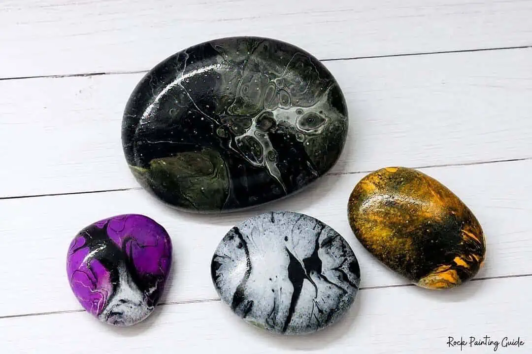 Halloween Paint Pouring on Rocks: Easy Acrylic Pour Tutorial 4 Halloween paint pouring rocks in black, purple, white, orange, and gold acrylic paint on a white wood background.