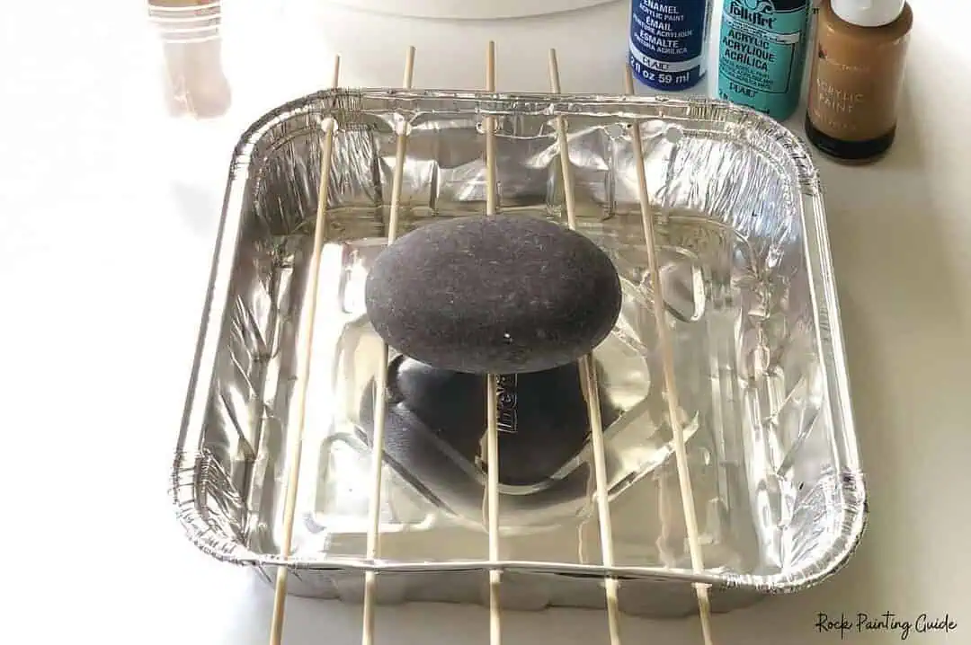Halloween Paint Pouring on Rocks: Easy Acrylic Pour Tutorial 5 DIY paint pouring tray made from a foil baking pan and wooden skewers holding a smooth rock