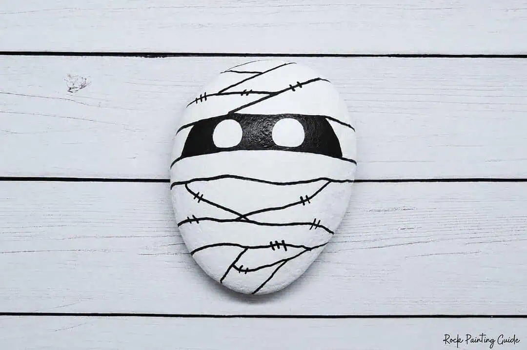 Easy Mummy Rock Painting: Step-by-Step Halloween Tutorial 7 Mummy-themed painted rock with black eyes and stitched bandages on white background.