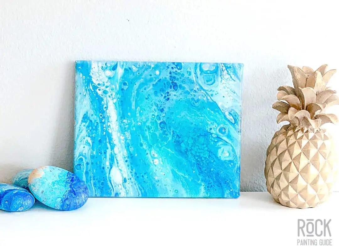 8 Easy Paint Pouring Techniques to Create Beautiful Wall Art 4 ocean paint pouring wall art displayed on a table with a gold pineapple and painted stones