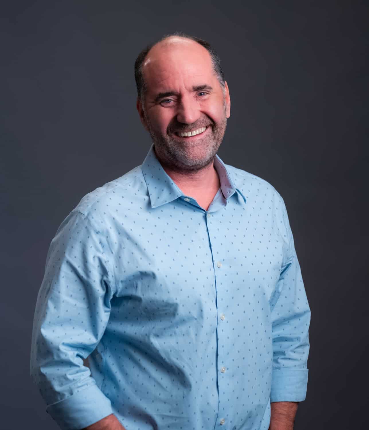 Man smiling, wearing a light blue patterned shirt, standing against a dark background.