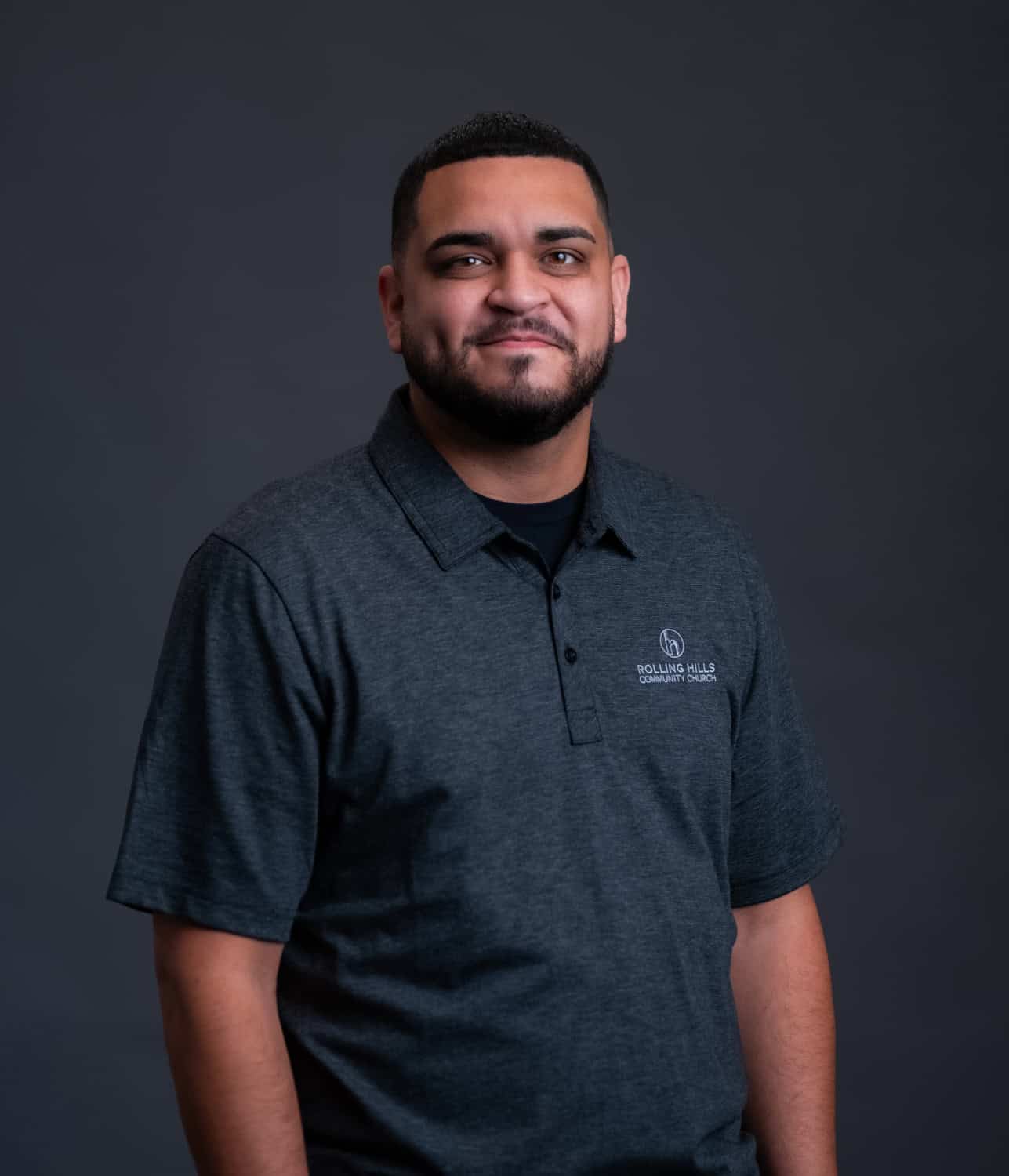 Community Church staff member in dark gray polo shirt with Rolling Hills logo, standing confidently against a plain dark background.