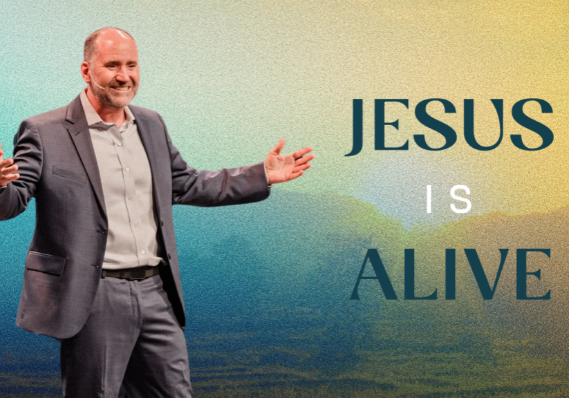 Man in suit with open arms, smiling, with mountains in background, promoting Jesus' resurrection.