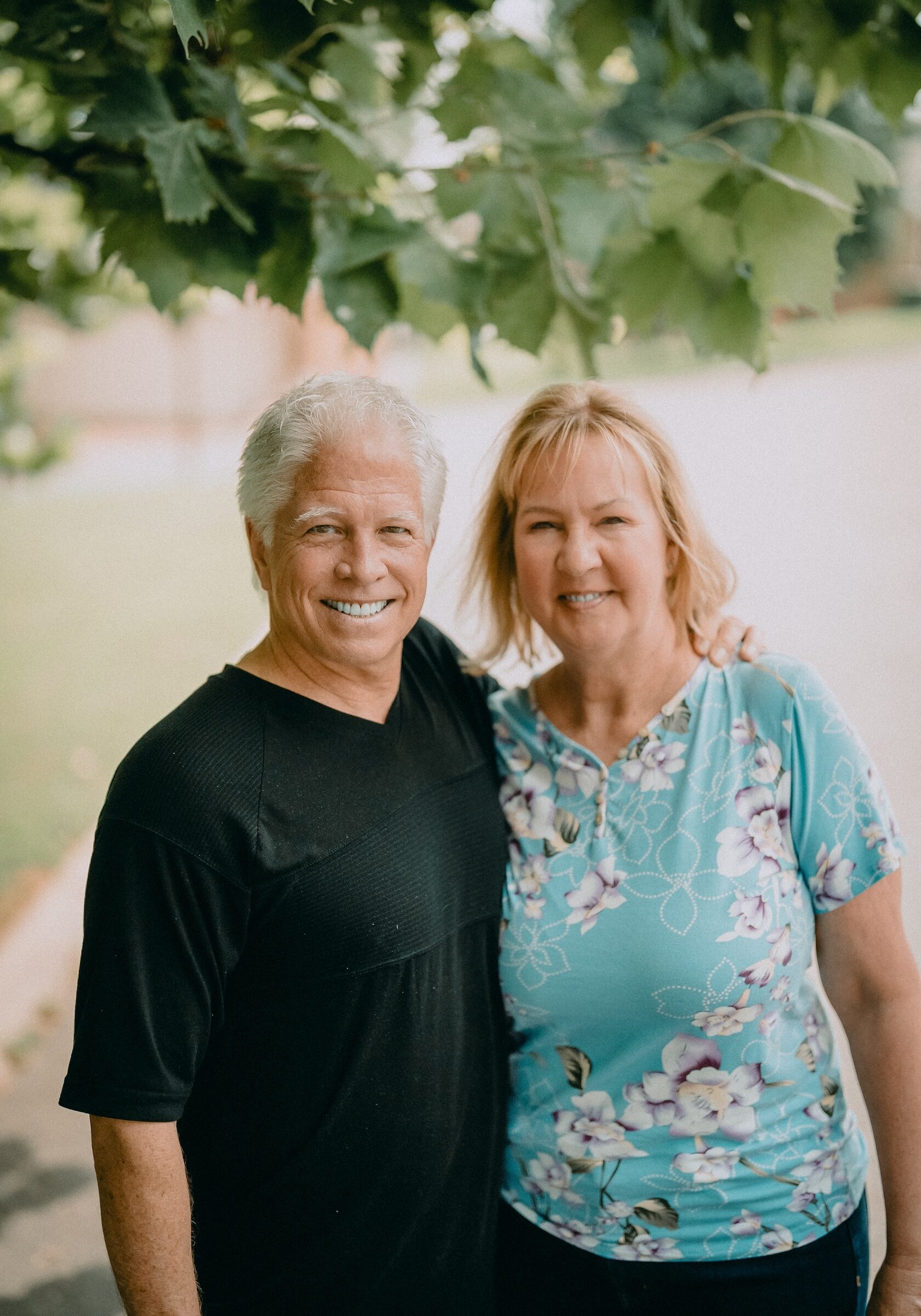 Older man and woman smiling under a leafy tree, embracing each other.