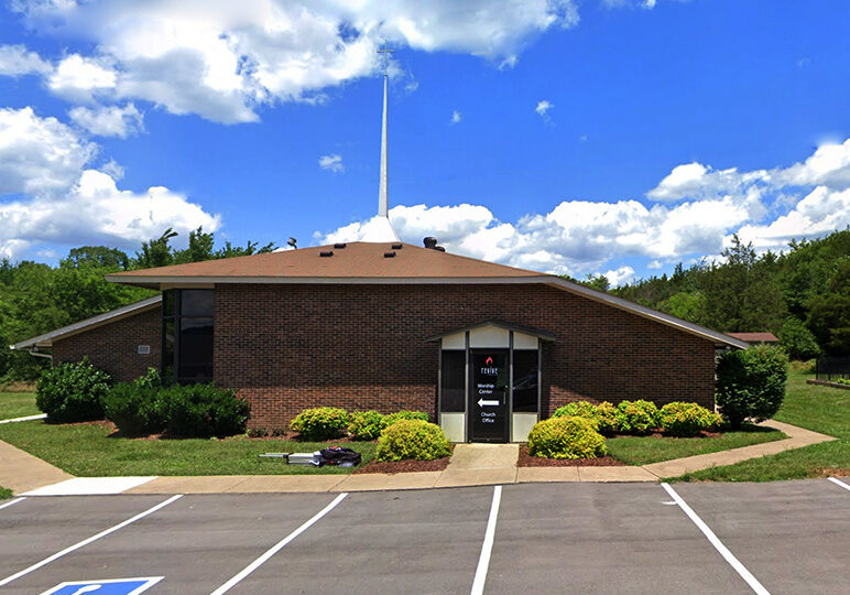 Modern brick church building with a steeple and parking lot, surrounded by greenery under a blue sky.