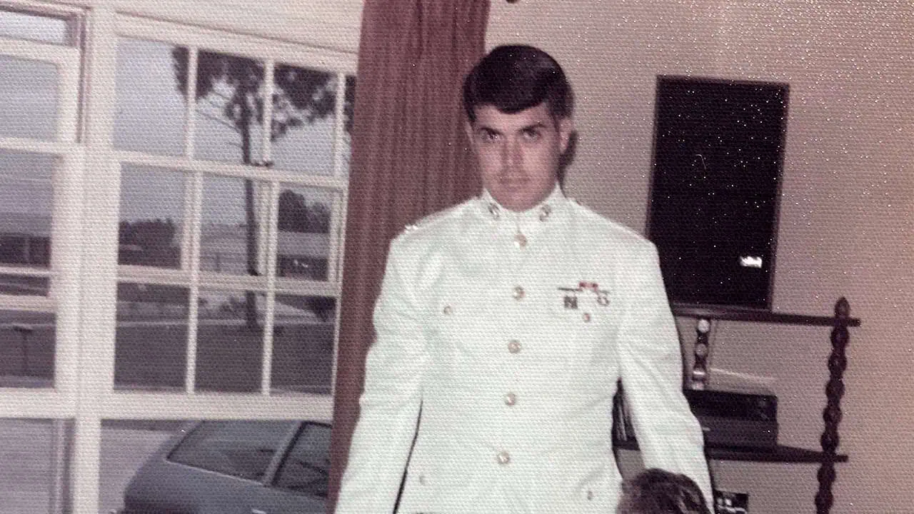 Young man in a white military uniform with medals, indoors near a window.