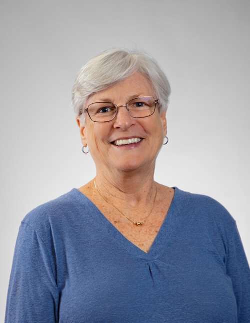 Smiling senior woman in blue top with glasses against plain background.
