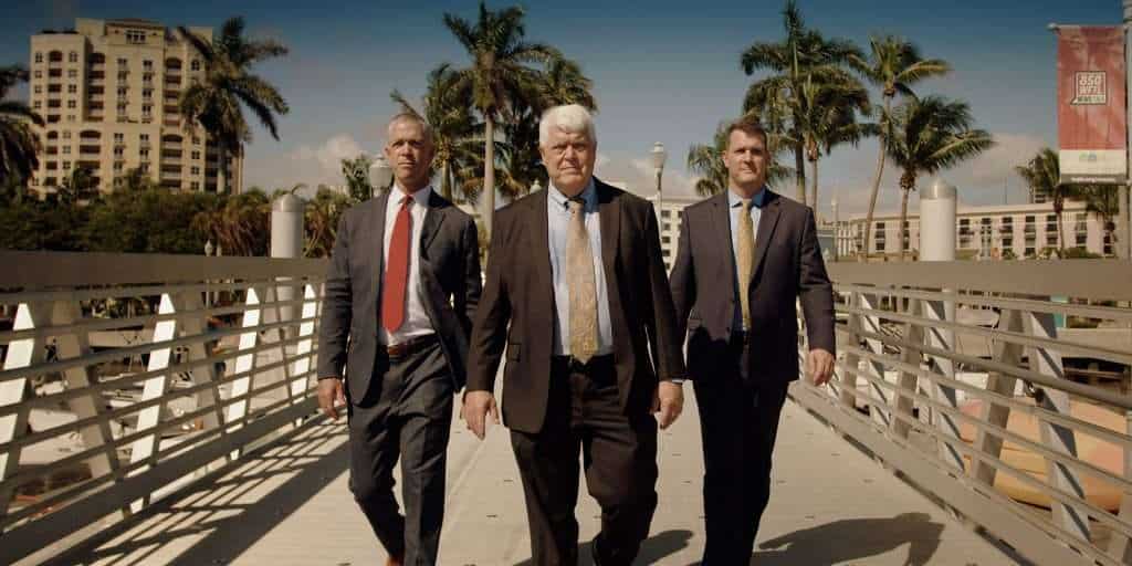 Three professional men walking on a bridge in a city with palm trees and buildings in the background.
