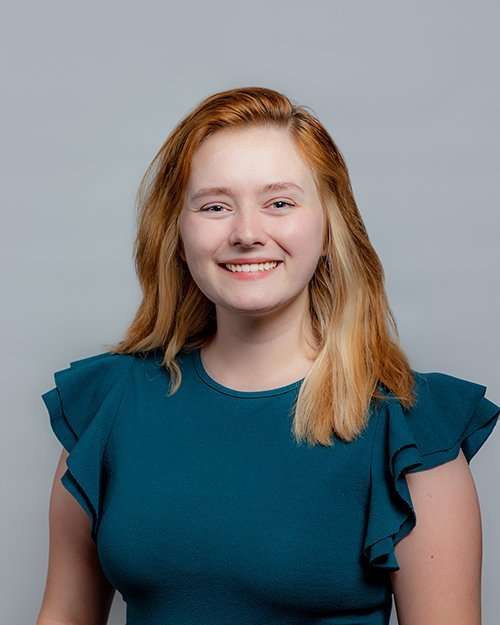 Smiling young woman with red hair in a blue top against a gray background.