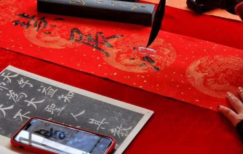 Traditional Lunar New Year craft of writing spring couplets with a brush on red paper, featuring intricate dragon patterns and ink tools