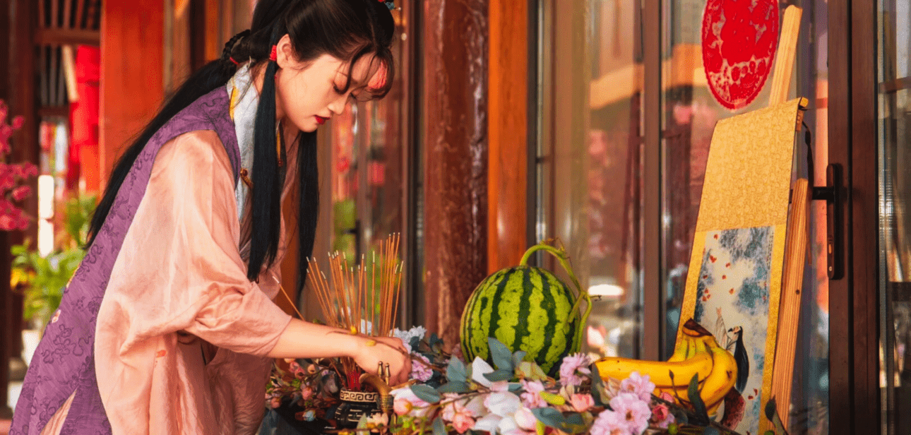 Woman lighting incense sticks at a modern ancestor worship altar with fruit and floral decorations.
