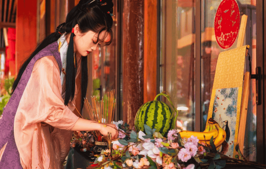 Woman lighting incense sticks at a modern ancestor worship altar with fruit and floral decorations.