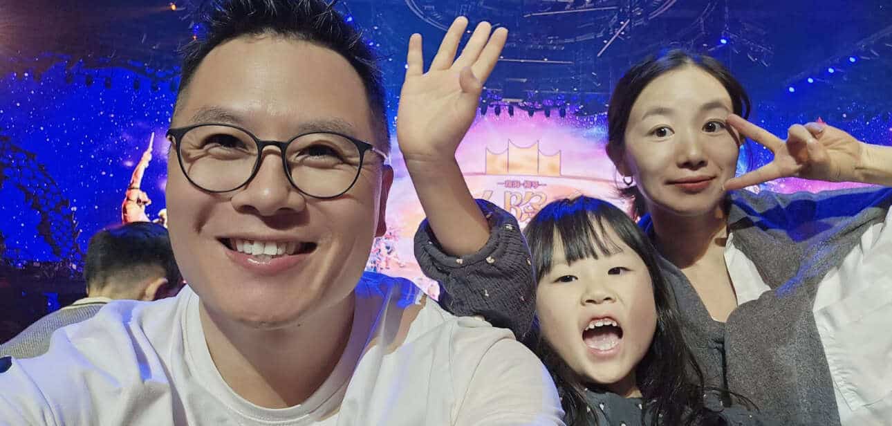 A cheerful family selfie with a father, mother, and young daughter at an event. The background features a colorful