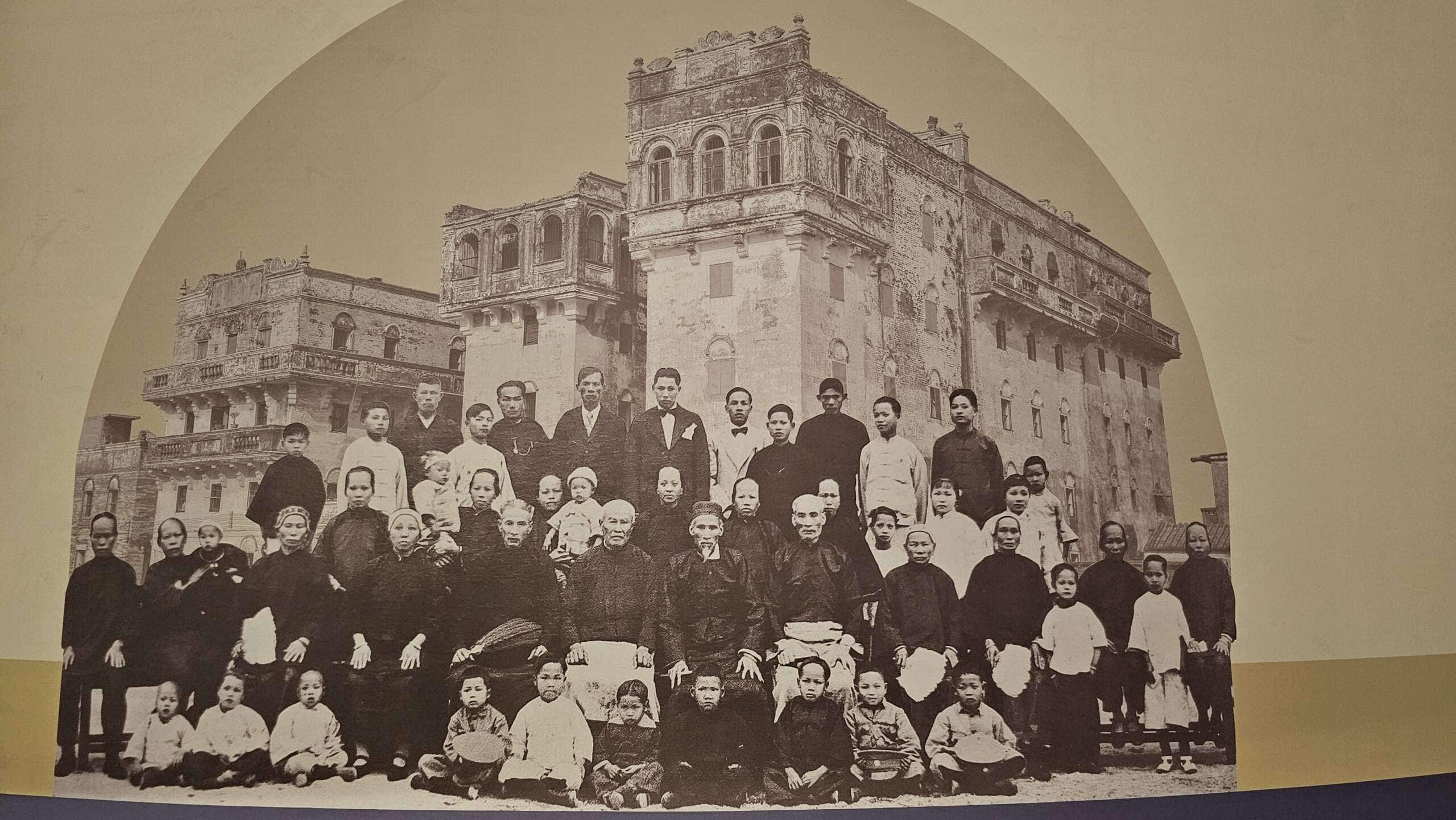 Historical photograph of a large Taishanese family in front of iconic diaolou towers, showcased at Taishan Overseas Chinese Culture Museum.
