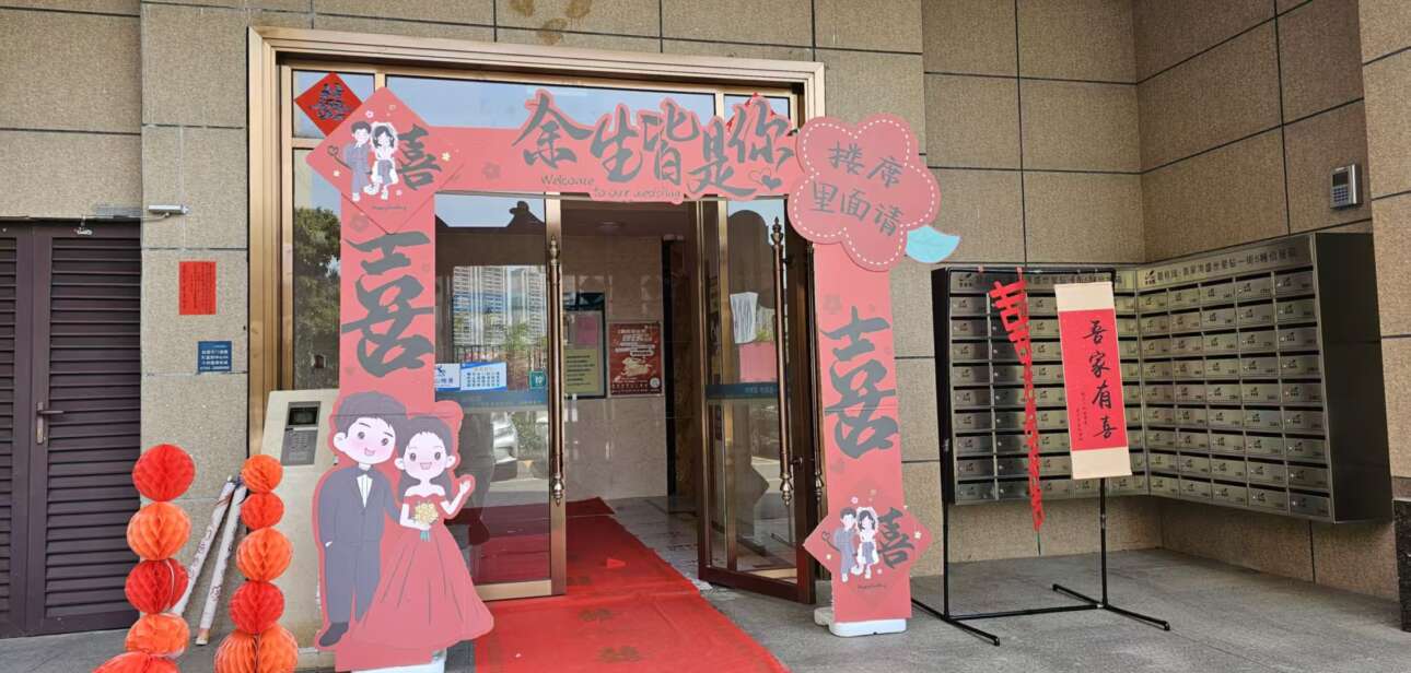 A wedding setup at a building entrance with a red carpet, cutouts of a bride and groom, and Chinese decorations, celebrating Guangdong’s marriage traditions.