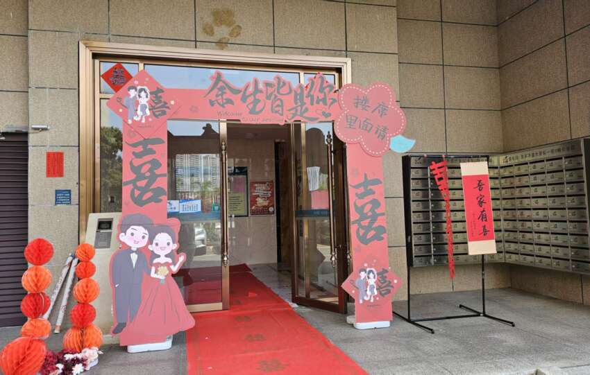 A wedding setup at a building entrance with a red carpet, cutouts of a bride and groom, and Chinese decorations, celebrating Guangdong’s marriage traditions.