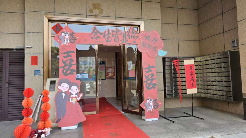 A wedding setup at a building entrance with a red carpet, cutouts of a bride and groom, and Chinese decorations, celebrating Guangdong’s marriage traditions.