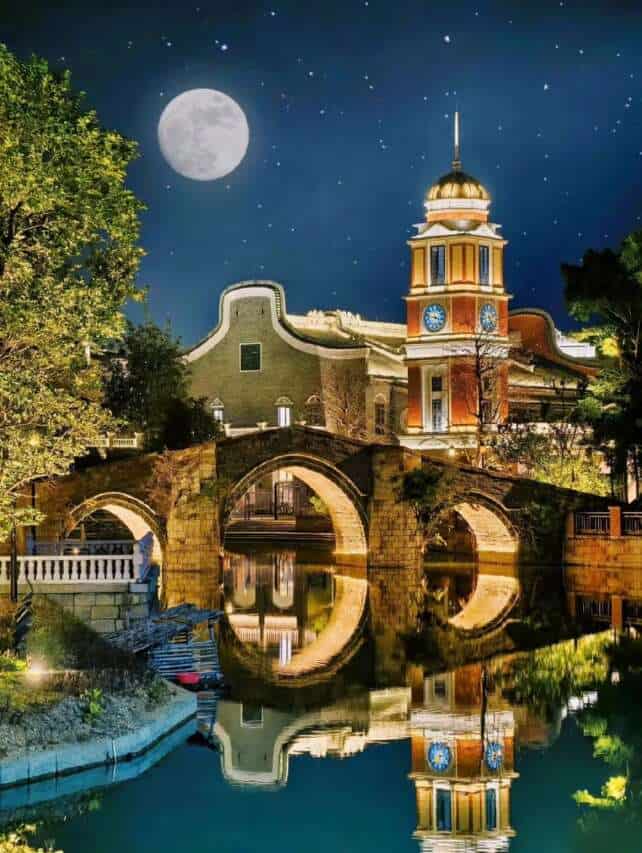 Chikan in Kaiping at night, featuring a stone bridge, a clock tower, and traditional buildings reflecting on the water under a full moon.