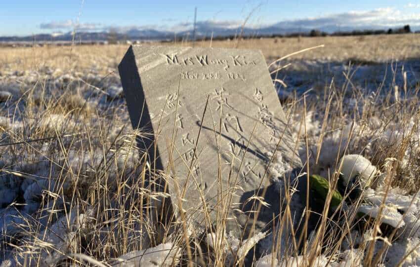A weathered grave marker bearing Chinese inscriptions stands amidst snow-covered fields, marking the resting place of Mrs. Wong Kim—a testament to the enduring legacy of Montana’s Chinese pioneers