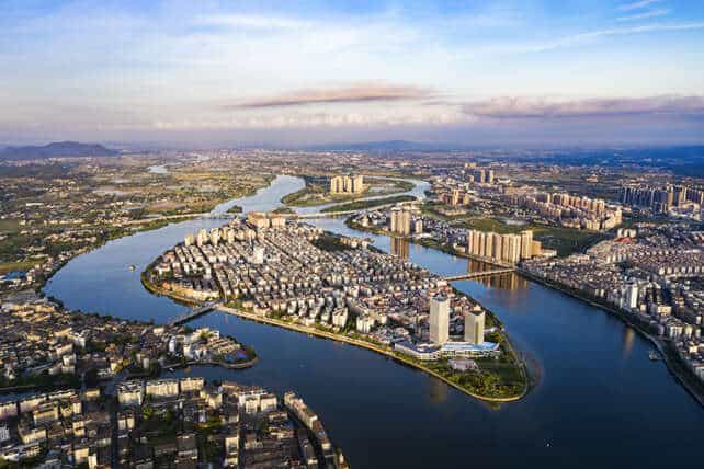 An aerial view of Kaiping in Guangdong Province, showing the Tan River cutting through the city with modern buildings, bridges, and urban infrastructure under a partly cloudy sky, surrounded by distant hills.