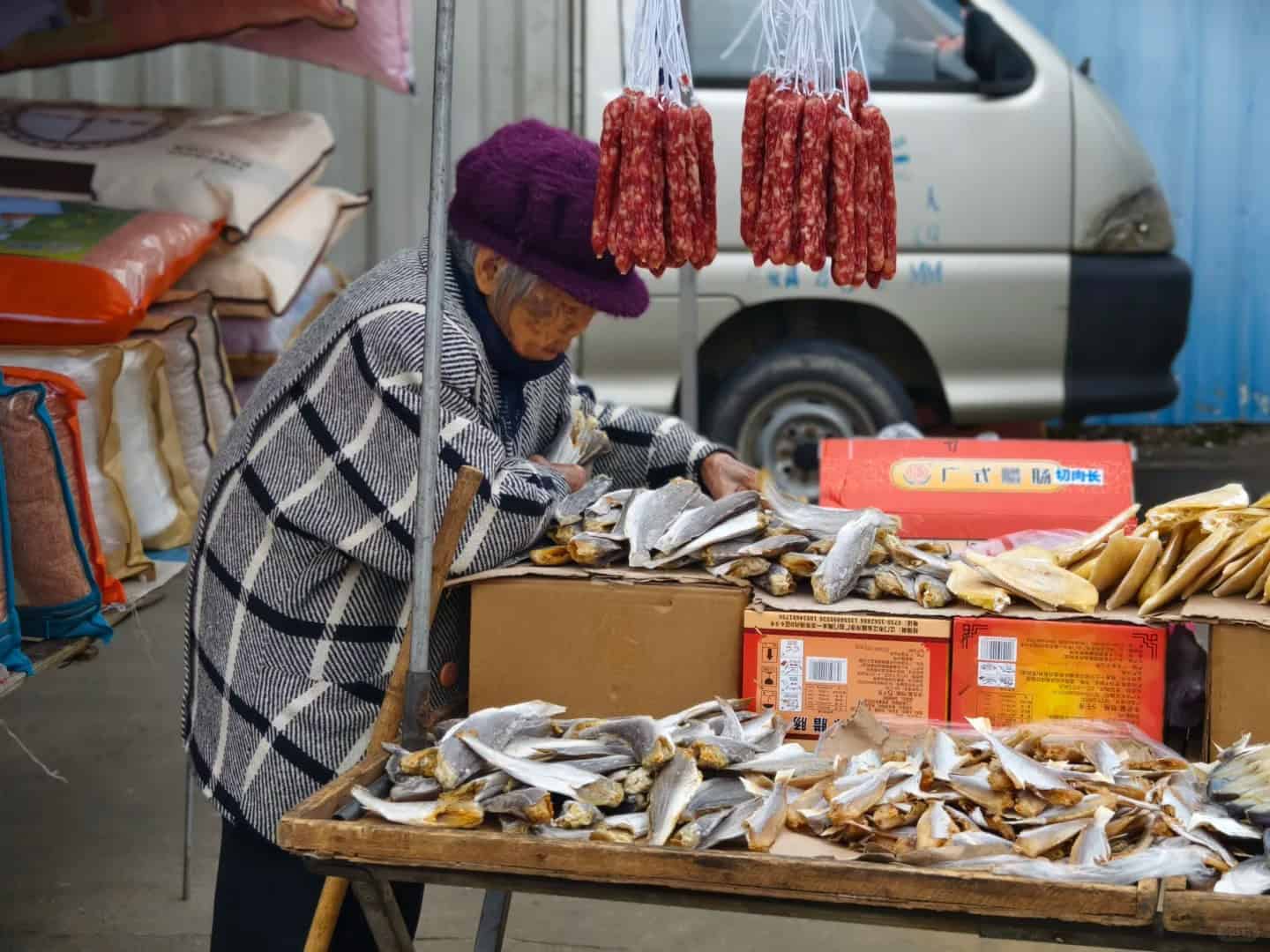 An elderly woman selling dried fish and sausages at a roadside market stall in a rural village in China.