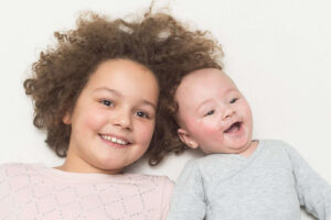 Lächelndes Geschwisterpaar liegt nebeneinander auf hellem Untergrund – ein älteres Kind mit lockigem Haar und ein fröhliches Baby in grauem Strampler.