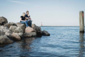 Familie mit Kleinkind sitzt gemeinsam auf großen Steinen am Meer, mit Blick auf einen Leuchtturm in der Ferne.