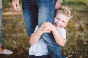 Lachender Junge umarmt liebevoll das Bein eines Erwachsenen im Freien.