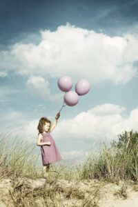 Kleines Mädchen mit drei violetten Luftballons steht barfuß auf einer Düne in Warnemünde unter blauem Himmel mit Wolken.