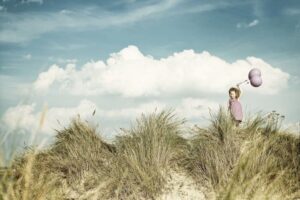 Kind mit violetten Luftballons steht auf einer Düne in Warnemünde vor blauem Himmel mit weißen Wolken.