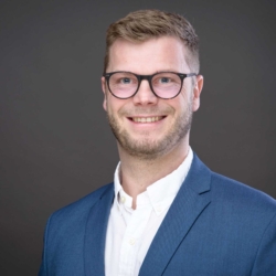 Bewerbungsfotos im Rostocker Fotostudio: Junger Mann mit Brille und blauem Anzug lächelt professionell vor einem dunklen, neutralen Hintergrund. Sein seriöses und sympathisches Auftreten vermittelt Kompetenz.