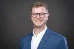 Bewerbungsfotos im Rostocker Fotostudio: Junger Mann mit Brille und blauem Anzug lächelt professionell vor einem dunklen, neutralen Hintergrund. Sein seriöses und sympathisches Auftreten vermittelt Kompetenz.