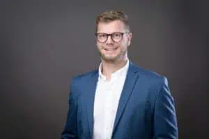 Bewerbungsfotos im Rostocker Fotostudio: Junger Mann mit Brille und blauem Anzug lächelt sympathisch vor einem dunklen, neutralen Hintergrund. Sein professionelles Erscheinungsbild vermittelt Kompetenz und Selbstbewusstsein.
