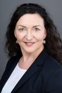 Bewerbungsfotos im Rostocker Fotostudio: Frau mit dunklen, lockigen Haaren in Business-Outfit lächelt selbstbewusst vor einem neutralen, dunklen Hintergrund. Ihr professionelles Auftreten vermittelt Kompetenz und Sympathie.