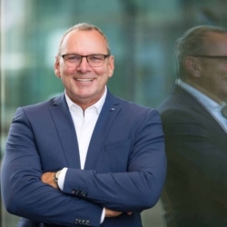 Bewerbungsfoto Outdoor vom Geschäftsmann im blauen Anzug mit Brille, lacht selbstbewusst vor einer modernen Glasfassade. Sein Spiegelbild ist im Hintergrund sichtbar.