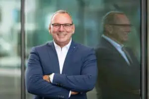 Bewerbungsfoto Outdoor vom Geschäftsmann im blauen Anzug mit Brille, lacht selbstbewusst vor einer modernen Glasfassade. Sein Spiegelbild ist im Hintergrund sichtbar.