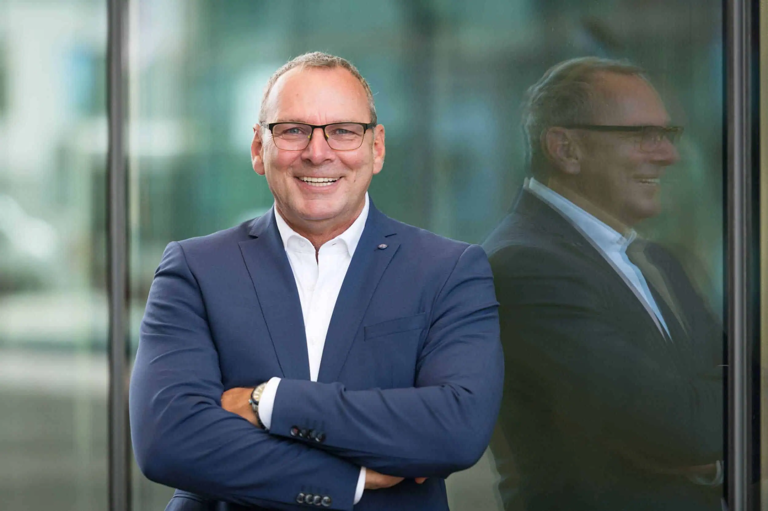 Professionelles Bewerbungsfoto Outdoor bei der MED in Rostock: seriös, sympathisch & modern Bewerbungsfoto Outdoor vom Geschäftsmann im blauen Anzug mit Brille, lacht selbstbewusst vor einer modernen Glasfassade. Sein Spiegelbild ist im Hintergrund sichtbar.